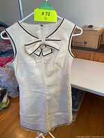 Front view of white sleeveless mini dress with navy trim and bow detail on chest, hanging on white hanger with yellow lot tag.