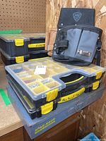Five hardware totes stacked including resin plastic totes with clear lids and one metal case, with two fabric tool belts leaning on top