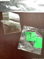 Two small clear plastic bags lying on a wooden surface, one containing a small square gold-toned metal piece, the other empty or containing a small fragment.