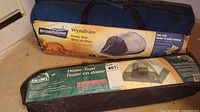Two dome tents in bags on floor; Broadstone Wyndham 6 person tent in blue bag with yellow label, Escort 3 person dome tent in black bag with green label.