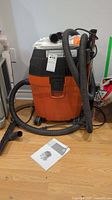 Front view of FEIN Turbo II HEPA wet dry vacuum in orange and black with hose wrapped around and user manual on floor.