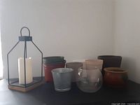 Photo showing black metal lantern with wooden base and white battery-operated candle alongside eight assorted planters in various sizes, materials and colors. Set against plain off-white wall.