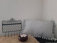 Photo showing robin egg blue decorative pillow with fringed tassel edges, black metal jewelry hanger with scroll pattern and wire grid, and round vintage ceramic trinket box with rose lid on a textured surface.