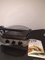 Stainless steel electric grill with black plastic accents, hinged top lid, three control knobs on front, detachable grease drip tray underneath, placed on a black surface with instruction manual beside it.