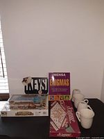 Photo showing Mensa Enigmas puzzle book, vintage Scrabble board game, brand new set of four Stokes white ceramic cups, and London History Puzzle box.