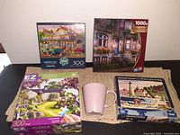 Front view of four boxed jigsaw puzzles and pink Ikea cup on a textured beige cloth.