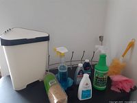 Ikea white plastic corner trash bin with dark lid and assorted cleaning supplies including spray bottles and brushes.
