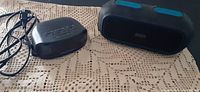 I-Home black waterproof Bluetooth speaker with blue accents and magnasonic alarm clock radio with cords on lace tablecloth.