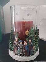 Glass hurricane candle holder with etched gold stars and detailed Christmas caroler base with six figures and trees, holding a large red pillar candle inside.