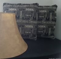 Photo showing two large square pillows printed with city names and part of the faux leather lamp shade.