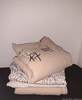 Stack of three folded queen-sized bedding items: beige duvet cover with black print on top, floral print sheet in middle, and solid beige flannel sheet at bottom.