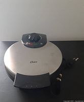 Closed view of Oster waffle maker showing stainless steel finish, handle, temperature dial and power cord.