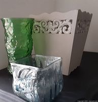 Large wooden planter with scalloped top edge and swirly cut-out pattern placed behind two glass vases, one green and one light blue/gray.