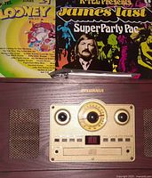 Front control panel of Sylvania Nostalgia turntable system with radio dial, speakers, cassette slot, and control buttons. Two vintage records visible behind the unit.