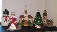 Group photo of all four ceramic Christmas village pieces arranged in a line, showcasing the church, lighthouse, Mrs. Claus, and snowman.
