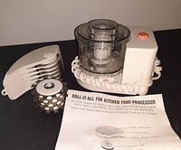 Mini food processor with coiled power cord, clear bowl, lid, white base with orange button, various plastic attachments, and original printed instructions laid out in front.
