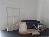 Lot items displayed on table including white wooden window planter frame with four glass panes, black planter box, three wood frames, metal watering can with flowers, and clear vase.