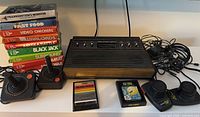 Atari 2600 console with two joystick controllers, two paddle controllers, and stacked game boxes with cartridges displayed in front.