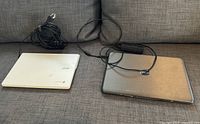 Both laptops shown closed with power cables on grey couch; Dell has visible paint spots on lid.