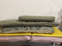 Stack of cushions including green fabric cushions, palm leaf patterned cushion, yellow cushions, and purple cushions underneath.