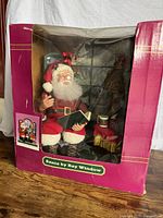Photo of boxed vintage automated Santa figurine seated by a bay window with lamp and presents, as seen from front.