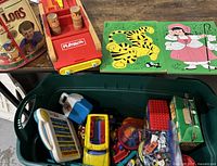 Bin of assorted toys including Lincoln Logs in a round container, wooden block puzzles with animal and character designs, red Playskool wooden bowling game, plastic toy cars, and Little People toys along with some plush toys.
