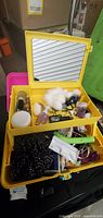 Open yellow plastic jewelry box showing assorted makeup, cotton balls, nail polish bottles, black beaded necklace, rings, hair clips, and other small items inside with fold-up mirror.
