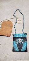 Two small crossbody purses on the ground, one turquoise with skull pattern and chain, the other black with beige suede pouch and gold chain.