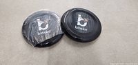 Photo showing two black Binske frisbees on a flat surface, one still in plastic wrap and the other unwrapped.