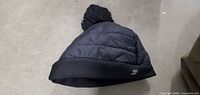 Black quilted winter cap with pom-pom on top and logo on cuff