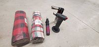 Photo showing two cross hatch patterned fabric rolls and three blow torches arranged on concrete.