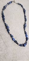 Photo of one blue beaded necklace featuring various shades and shapes of blue stones with silver spacer beads and a clasp closure on a light gray surface.