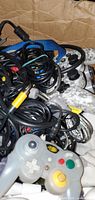 Lot of wired game controllers in cardboard box showing cables and assorted controller types including Xbox, PlayStation, and Nintendo GameCube.