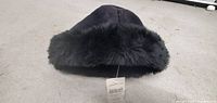 Black puffy winter hat with faux fur trim, shown from the front on grey carpet.
