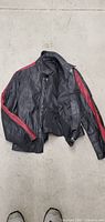 Black leather look women's coat with red stripes down the sleeves laid flat on a concrete floor, showing front and sleeves.