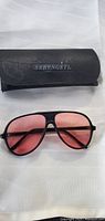 Black Serengeti branded protective case and sunglasses with black frame and pink lenses placed on a white surface.