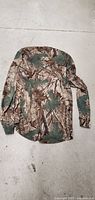 Back view of the long sleeve women's camo shirt laid flat on a concrete floor showing full camouflage pattern.