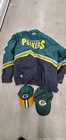 Front view of the vintage Green Bay Packers jacket with two caps on floor