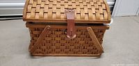 Woven picnic basket shown from front with leather strap latch and wooden handles folded down.