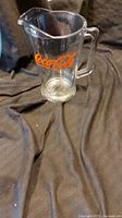 Clear glass Coca-Cola pitcher with orange logo shown on black cloth background