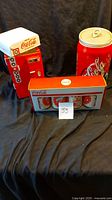 Photo showing two Coca Cola cookie jars, one shaped like a vintage vending machine with white top and one shaped like a soda can, along with boxed set of three Coca Cola glasses.