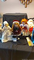 Photo shows three porcelain dolls prominently placed against a black cloth background. One doll is in white dress and bonnet, another in red Christmas-themed dress with Santa hat, and the third with curly red hair in Victorian-style dress. Visible also are several stuffed animals including teddy bears and a white plush duck.