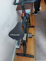 Angle view showing padded Raleigh seat cushion on black stationary exercise bike frame with foot pedals and straps.
