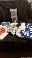 View of the hockey memorabilia lot including three hockey books, Maple Leafs wall clock, and white and blue Toronto Maple Leafs jersey, and a framed stamp picture.