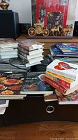 Photo showing stacked books including cookbooks with food images, coffee table books, DVDs, and a small decorative horse and carriage on a black surface.