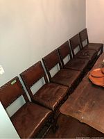 Six vintage dining chairs lined up against wall with brown leather upholstery, showing wear and scratches, with wooden frames and nailhead trim