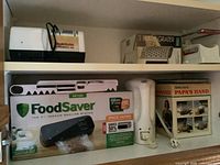 Shelf with boxed FoodSaver vacuum sealing system, white mandolin slicer, box grater, and other kitchen tools.