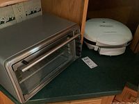 Photo showing Hamilton Beach stainless steel toaster oven with glass door, metal handle, and side control knobs on green countertop.