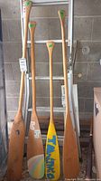 Four wooden paddles and one baseball bat leaning against a ladder inside a garage. Paddles vary in length and have painted blades or plain wood finish.