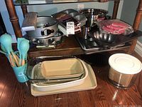 Overview of assortment of baking pans and kitchenware arranged on hardwood floor and chair rungs, showing pans, glass dishes, stainless steel bowl, utensil holder with turquoise utensils, and container with white lid.